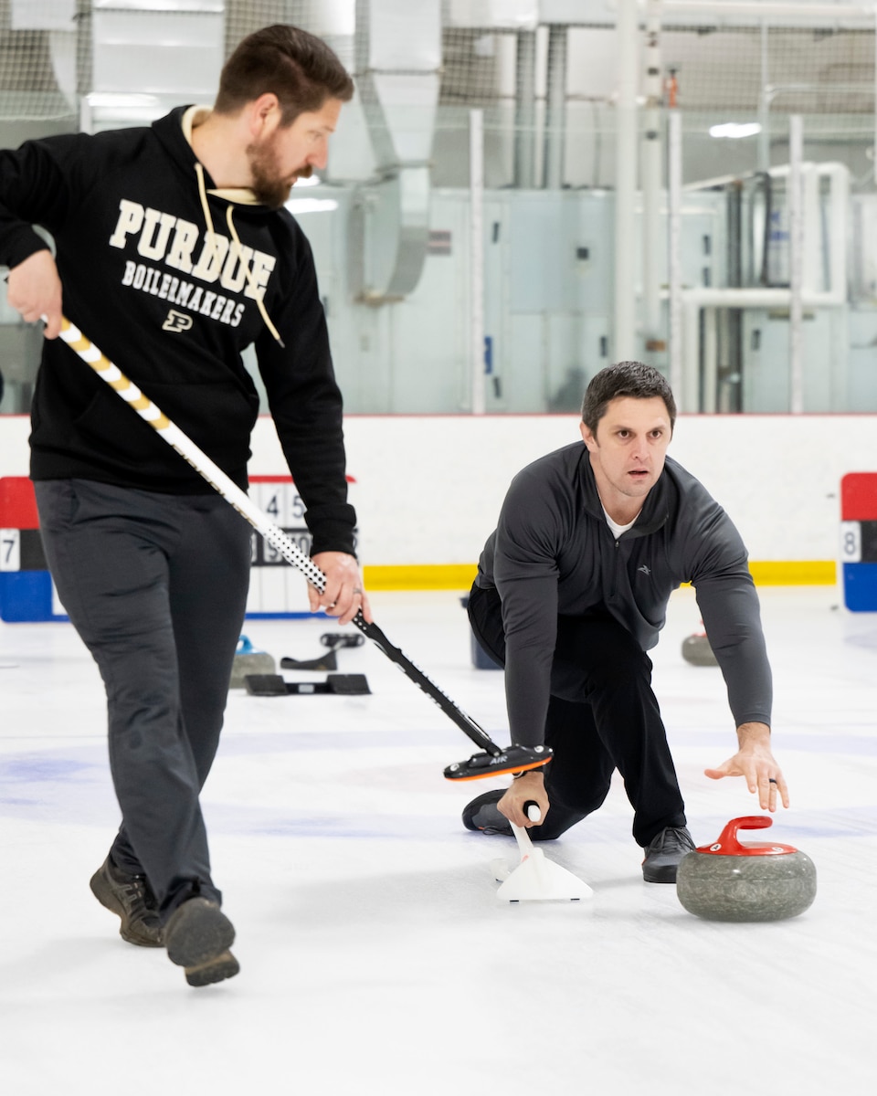 Two men, one standing, one kneeling, curl. The kneeling one, on the right, has just released a curling stone.