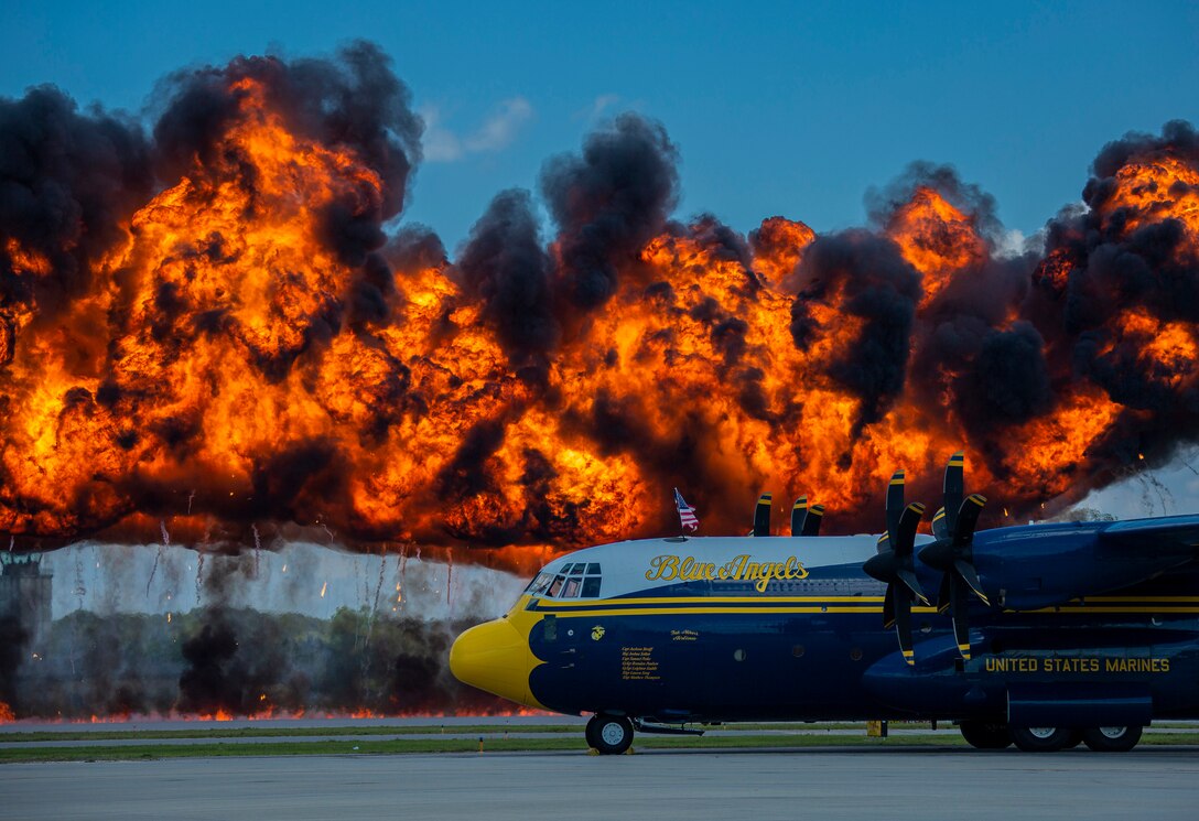 A Marine Corps aircraft is parked on the tarmac with fire behind it during an air show demonstration.