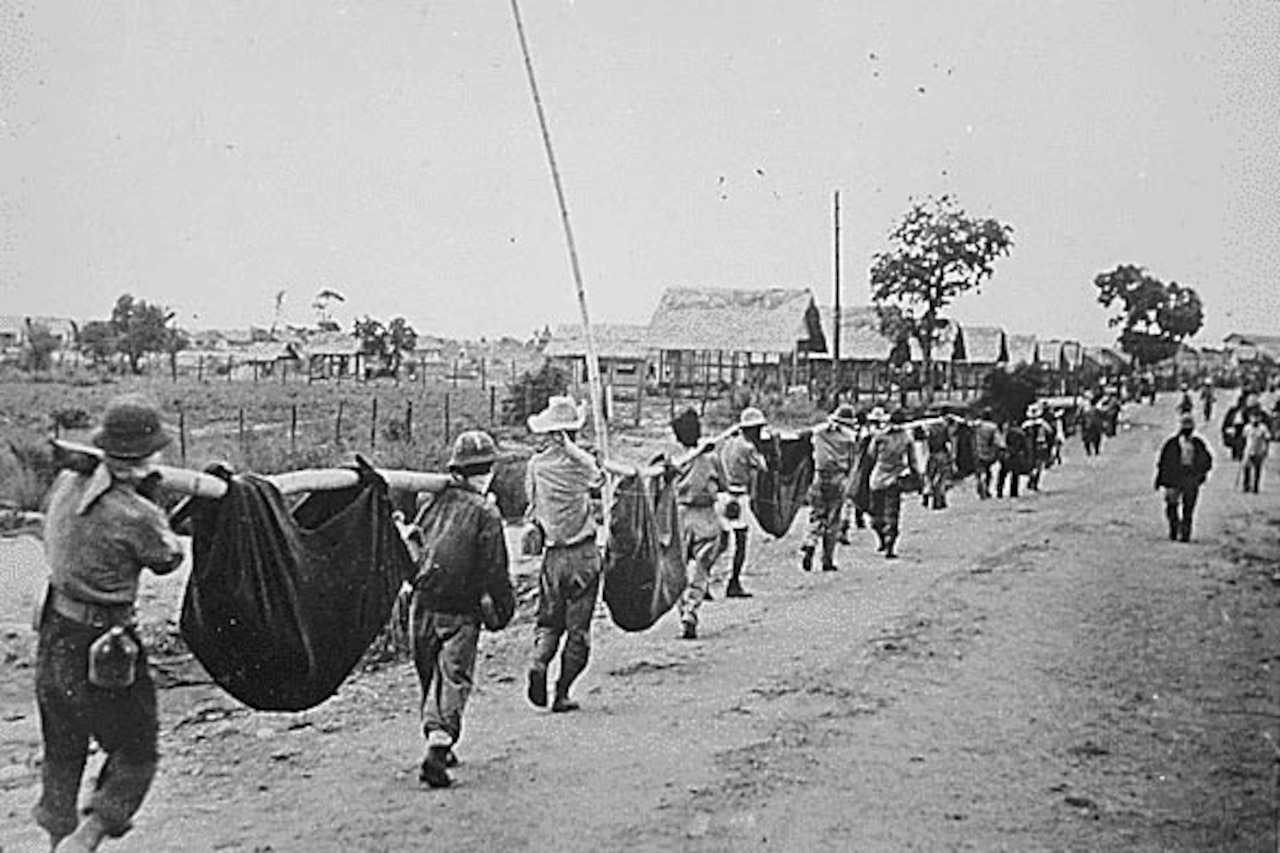 A line of men walks along a road. Many of the men carry improvised stretchers.