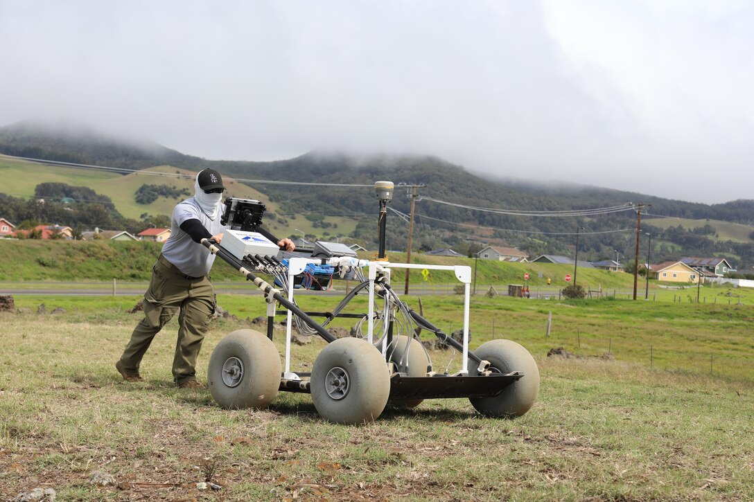 A man dressed in a gray T-shirt and long dark green pants leans in to push a piece of equipment across a pasture. The equipment rolls on four gray soft wheels.
