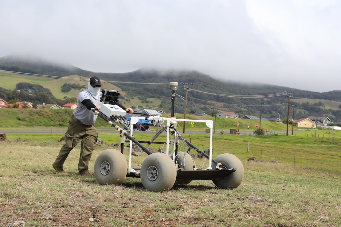 A man dressed in a gray T-shirt and long dark green pants leans in to push a piece of equipment across a pasture. The equipment rolls on four gray soft wheels.