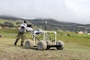 A man dressed in a gray T-shirt and long dark green pants leans in to push a piece of equipment across a pasture. The equipment rolls on four gray soft wheels.