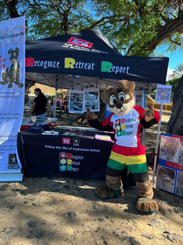 An adult is dressed in a dog mascot costume and standing on a beach. The dog is wearing a white t-shirt with a red, yellow and green stop light graphic that says RECOGNIZE, RETREAT, REPORT. The dog is standing to the right of an information booth with a black tent and black table cloth with the same stoplight image.