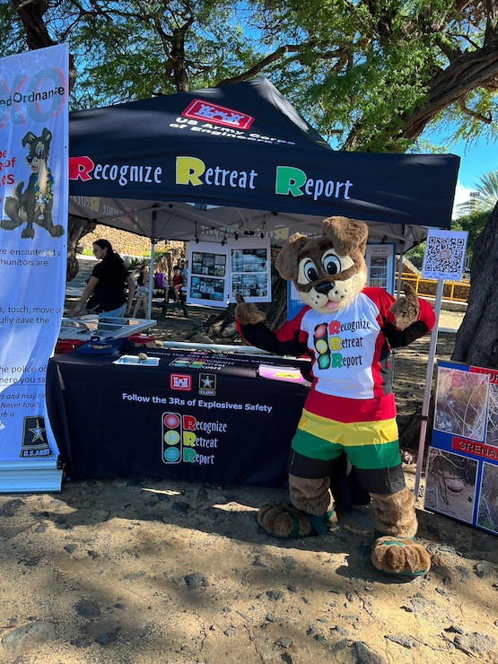 An adult is dressed in a dog mascot costume and standing on a beach. The dog is wearing a white t-shirt with a red, yellow and green stop light graphic that says RECOGNIZE, RETREAT, REPORT. The dog is standing to the right of an information booth with a black tent and black table cloth with the same stoplight image.