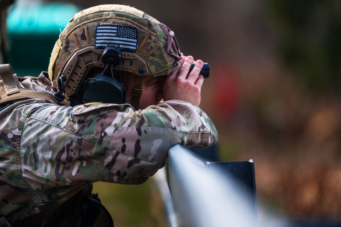 A soldier leans against a rail while looking through binoculars.