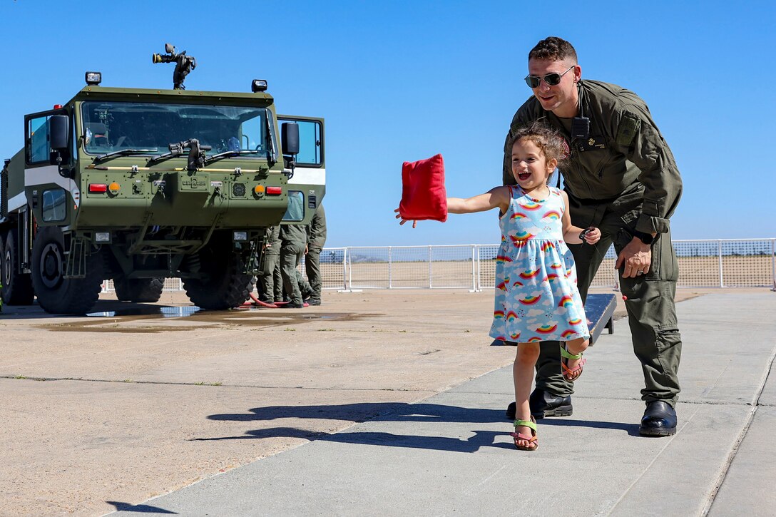 A child reaches to catch a small bean bag while a Marine smiles and looks on.