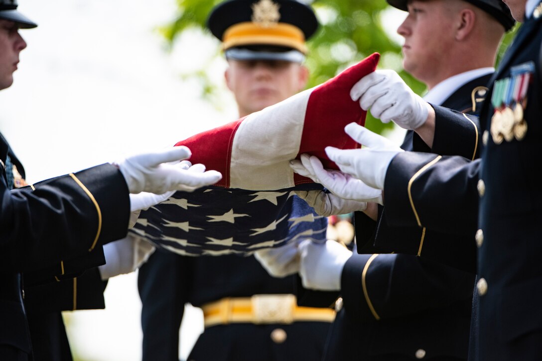 Soldiers in formal uniforms fold a flag during a funeral ceremony.