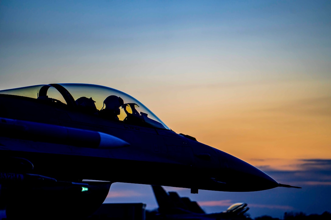 An airman shown in silhouette sits in the cockpit of a parked jet.