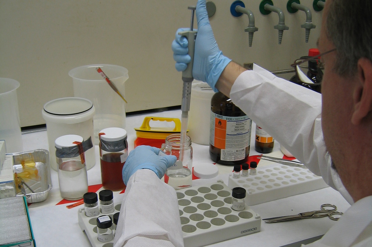 A man in a laboratory coat siting down uses a tool to extract liquid out of a glass jar.