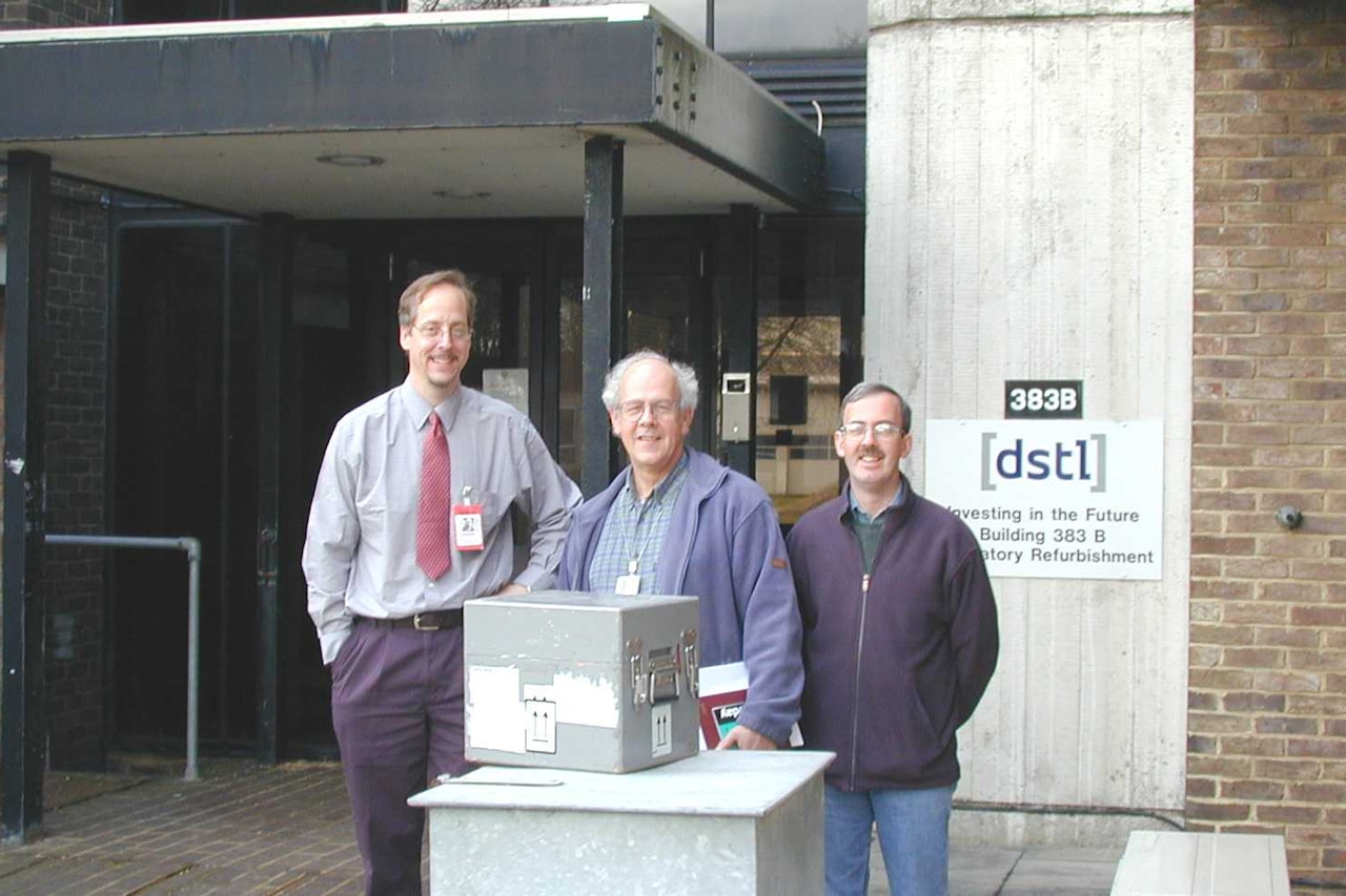 Three men pose in front of a container on a podium outside the entrance of a building.