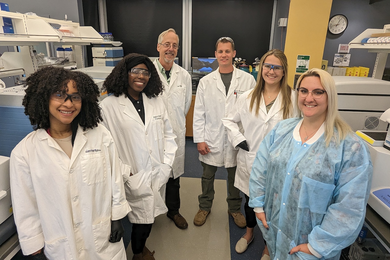 Six people in lab coats pose for a photo surrounded by lab equipment.