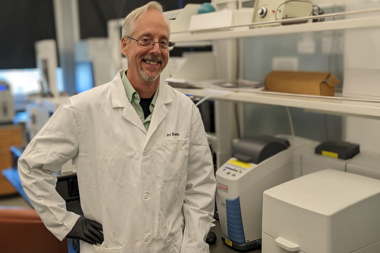 A man in a lab coat stands next to lab equipment.