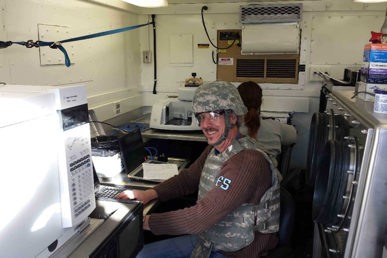 A man in flak vest and combat helmet smiles while sitting at a desk in a mobile lab.