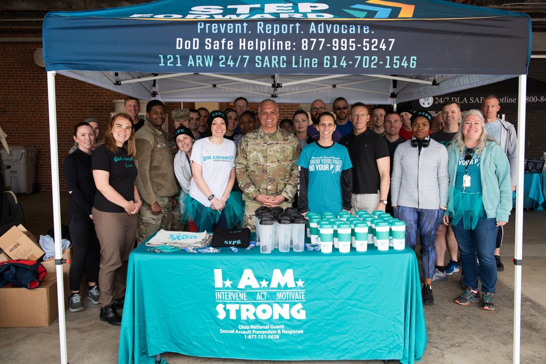 A large group of people standing in front of a teal SAPR booth with a teal tableclothed table and a booth tent which is also teal. A gray-haired man in the middle wears a military OCP camouflage uniform, the rest wear fitness attire.