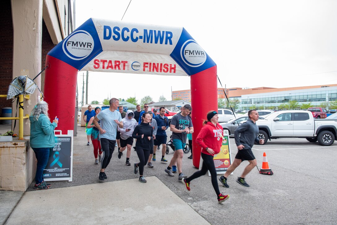 A gray-haired woman dressed in teal sounds the horn at the start of the run. Participants, men and women, in fitness attire run out of the gate. It is a inflatable res, white and blue gate.