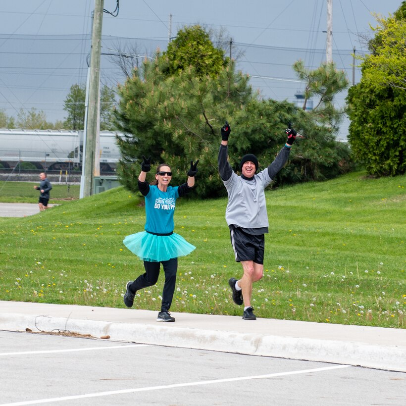 A dark-haired woman dressed in a teal T-shirt and wearing a tutu over her black jogging pants gives the peace sign as she runs alongside a man with a black winter hat on, gray shirt and black shorts who is also giving a peace sign with his fingers on a street next to the green grass of a golf course.