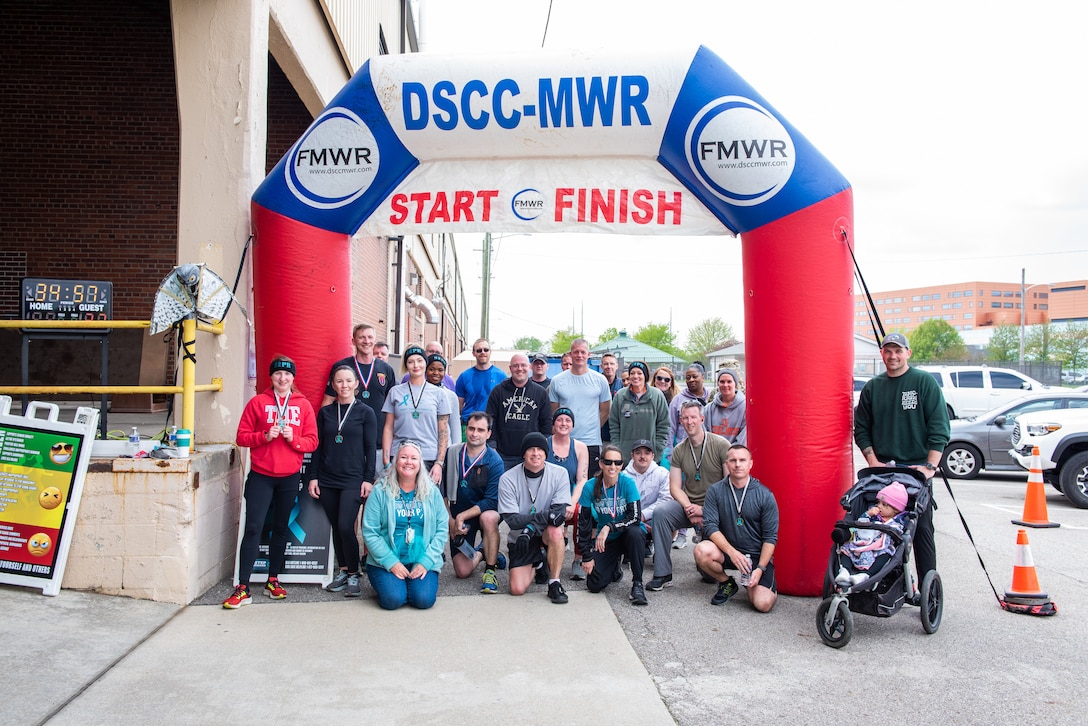 A large group of people standing in front of a inflatable race start/finish gate are smiling with medals after completing a race. The inflatable gate is red, white and blue. They are outside.