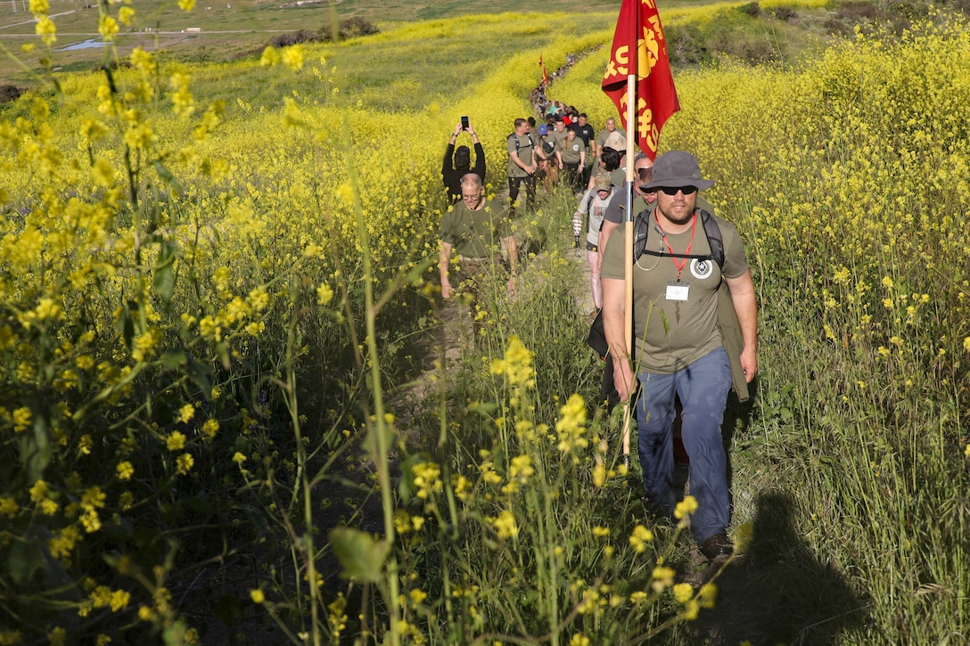 Retired and active service members carrying flags hike a field of tall grass.