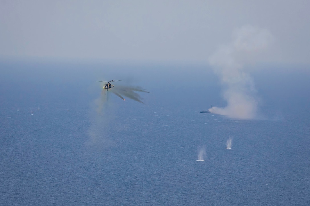 An airborne helicopter fires at a ship at sea leaving behind clouds of gray smoke.