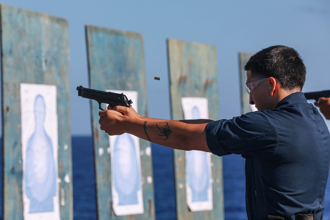 A sailor wearing eye protection glasses holds a M9 Beretta while aiming at a target.