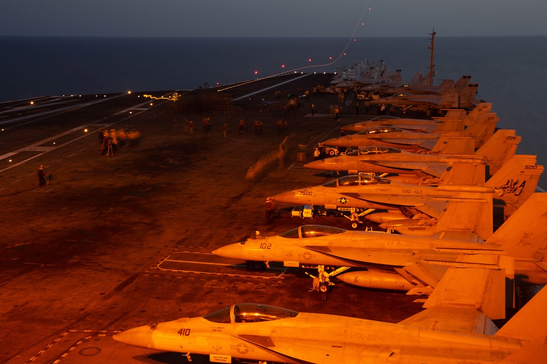 A row of planes sit on the flight deck of a ship while at sea.