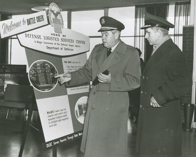 Two men stand in front of a sign with one man pointing at it.