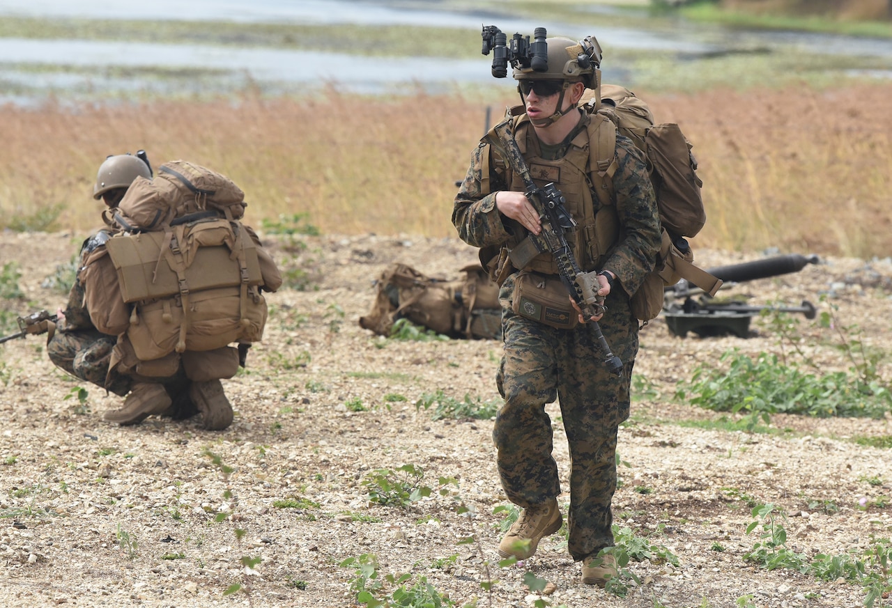 A service member moves across a field holding a weapon, as others look in different directions.