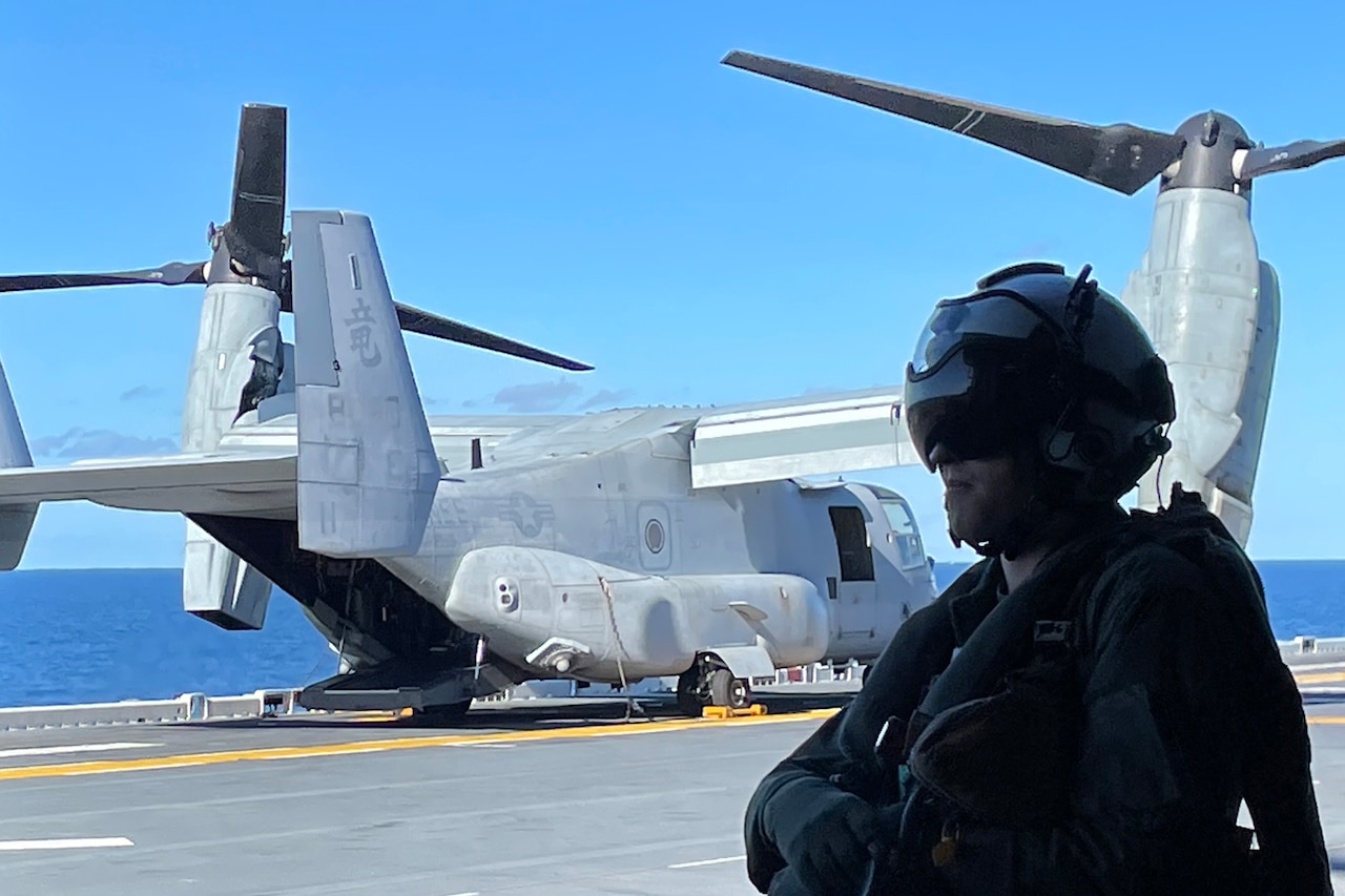 A service member stands in the foreground with a helicopter and the ocean behind him.