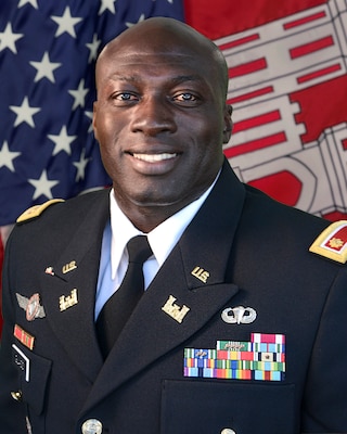 Black male is Army Dress Blues posing front of red, white and blue American Flag and the US Army Corps of Engineers castle flag, which is red and white.