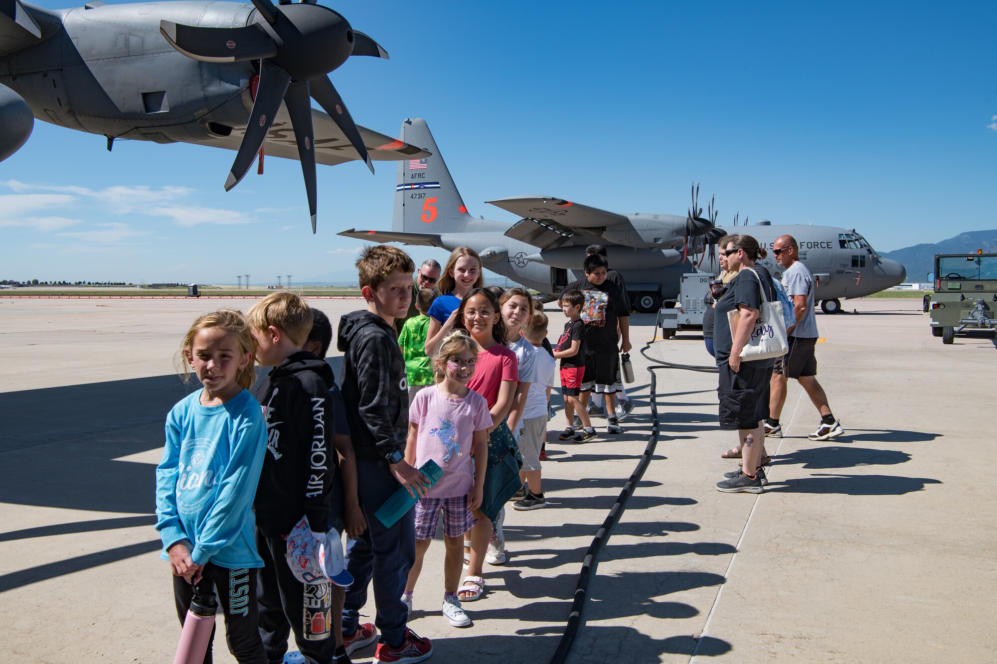 A group of children accompanied by chaperones line up in front of a C-130 with another C-130 in the background.