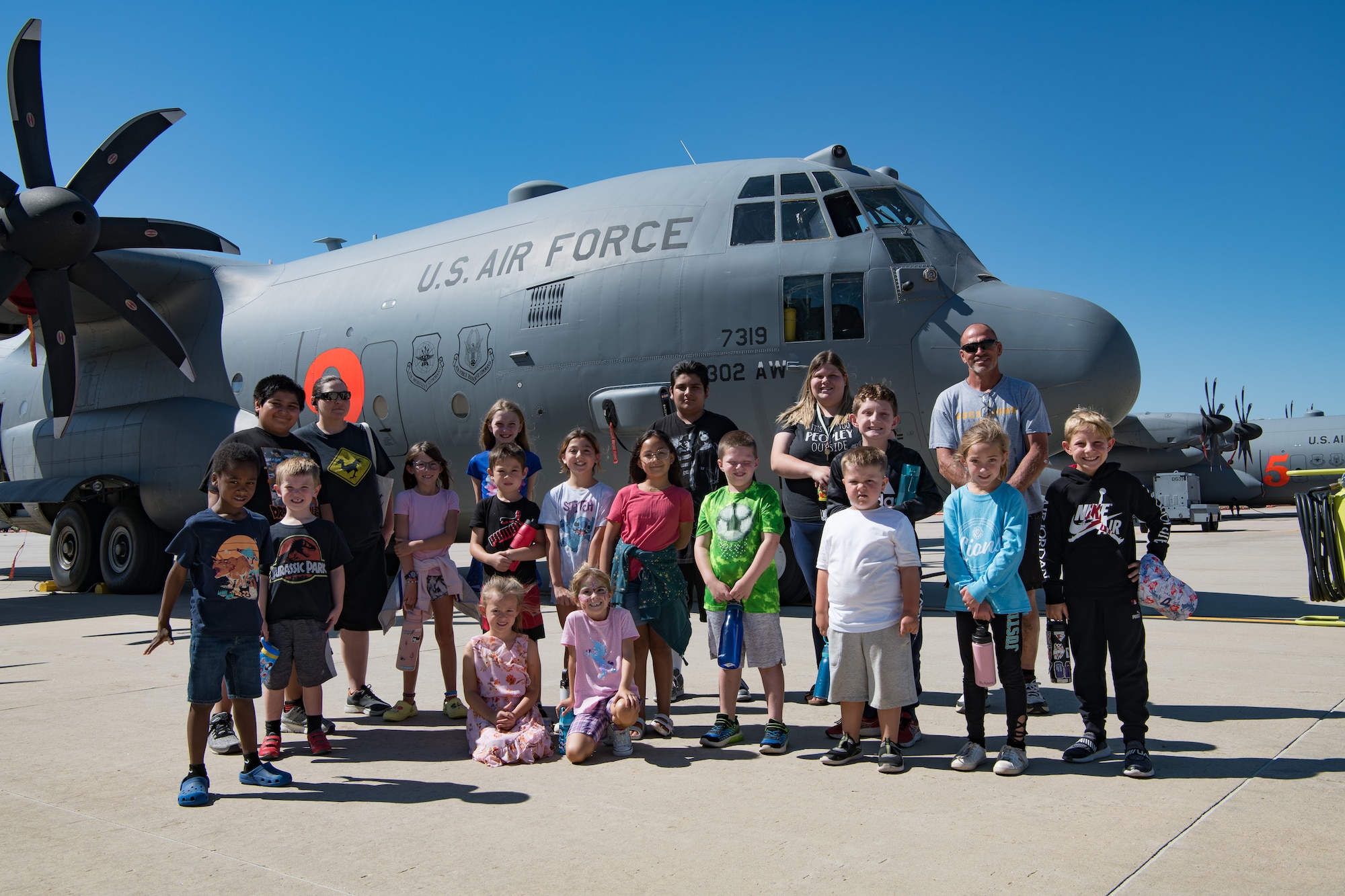 A group of children stand in front of a C-130 accompanied by chaperones in front of a C-130 aircraft.