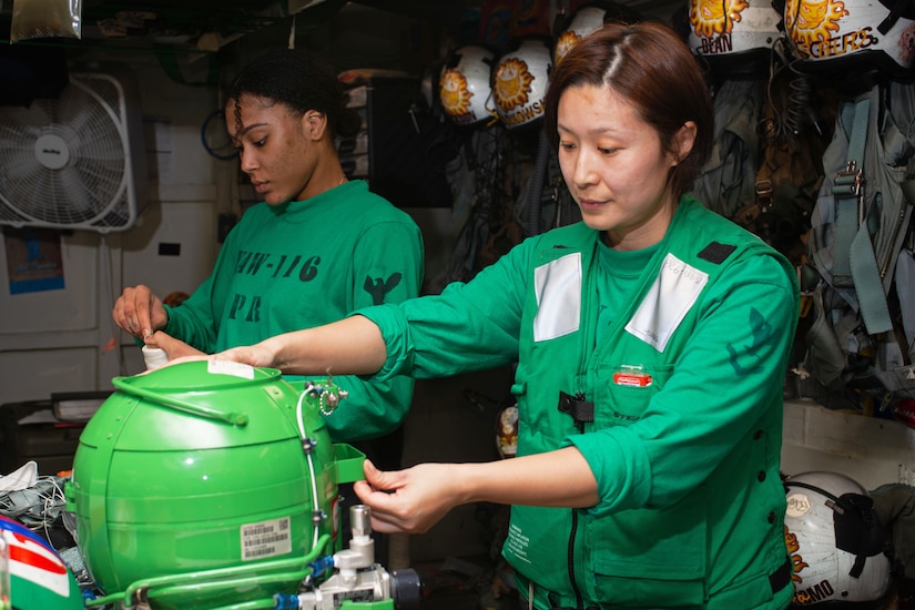 Two sailors in green uniforms work on oxygen equipment on the deck of a ship.