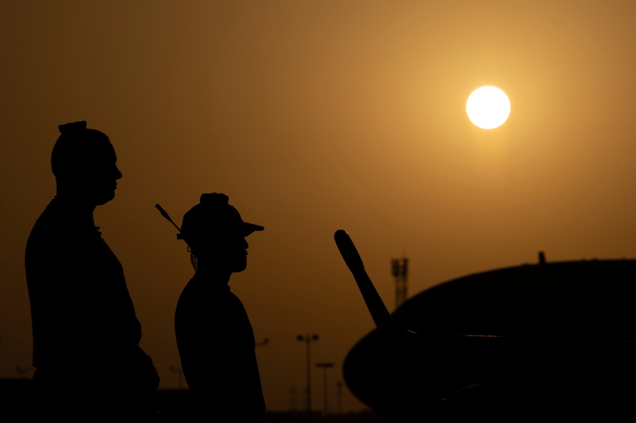 Two standing airmen and the nose of an aircraft are shown in silhouette against a dark-golden backdrop.