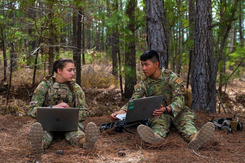Two airmen with laptops on their laps talk to each other while seated in a wooded area.
