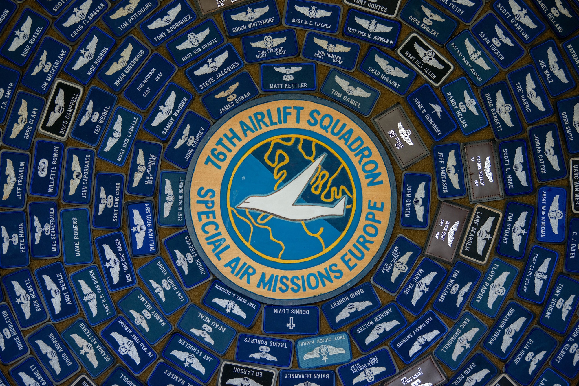 A board with patches from past members of the 76th Airlift Squadron is displayed on a wall