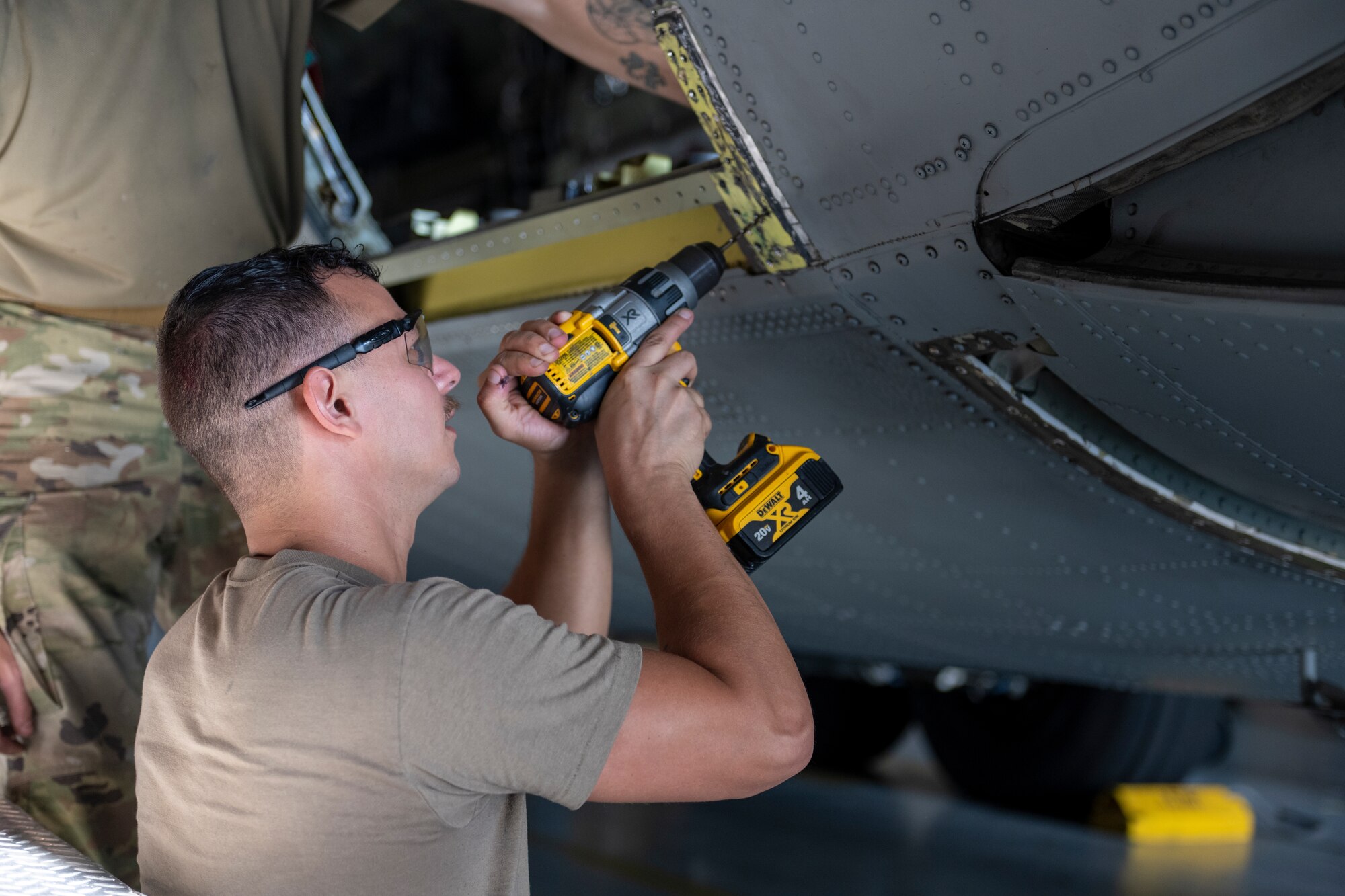 A man holding an electric drill, wearing safety glasses, drilling into the side of an aircraft.