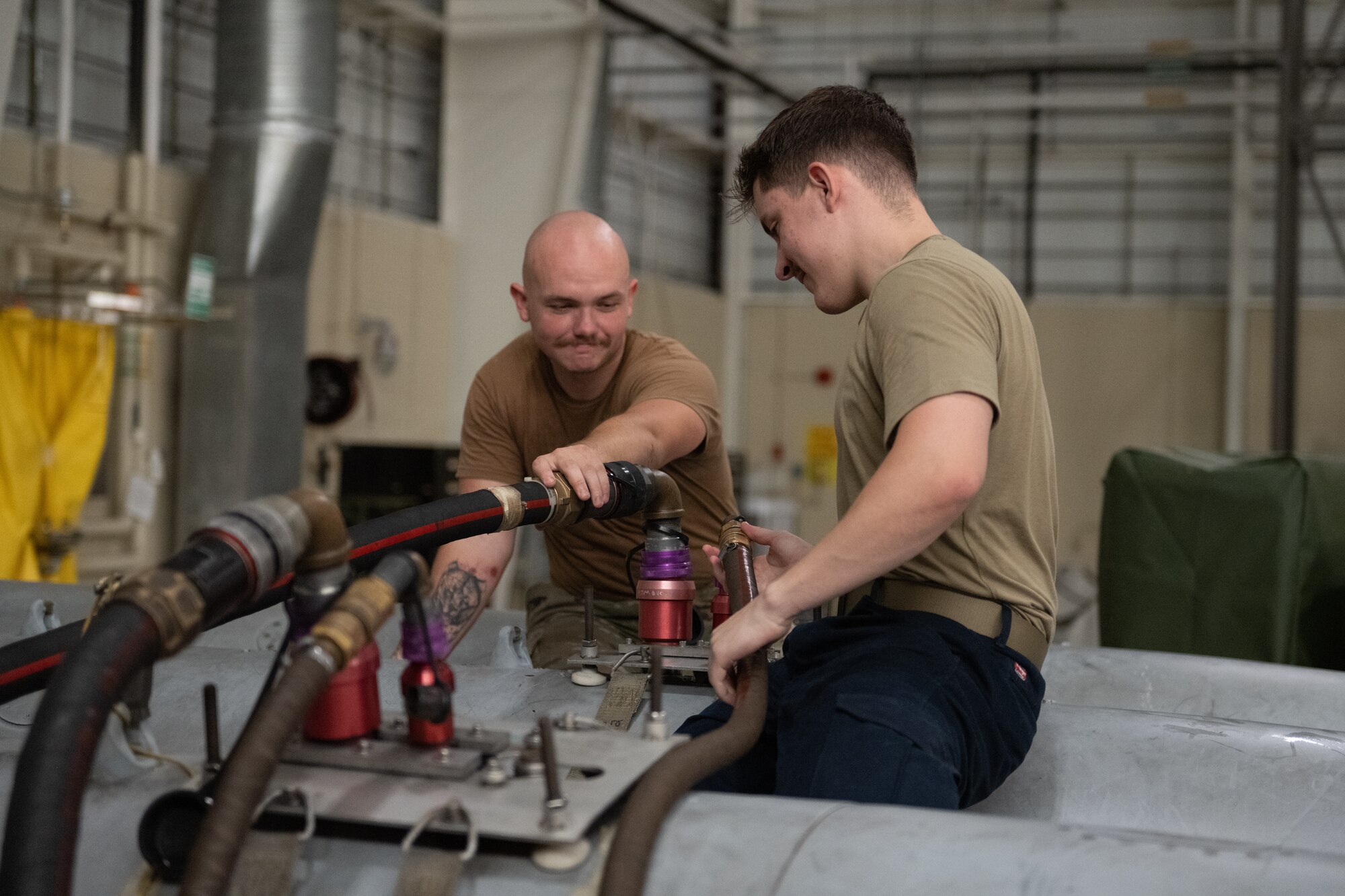 Two people sitting on top of grey cylindrical fuel tanks, holding onto a hose that is connected to the tanks.