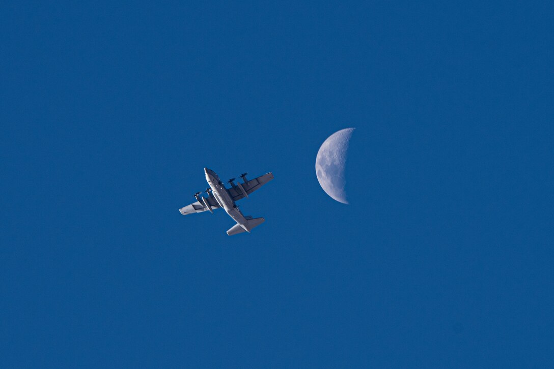 The underside of a military aircraft is shown flying during daylight with the moon behind it.