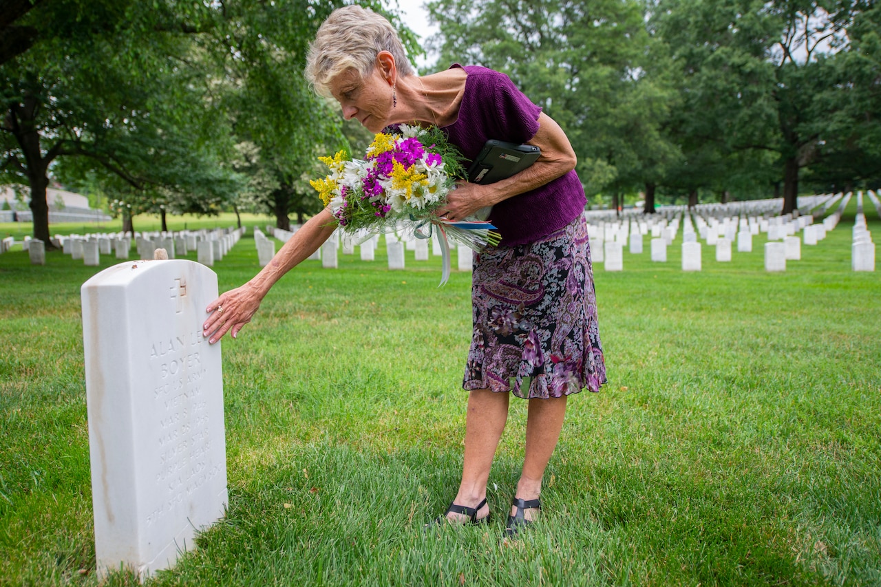 A woman holding flowers touches a gravestone.