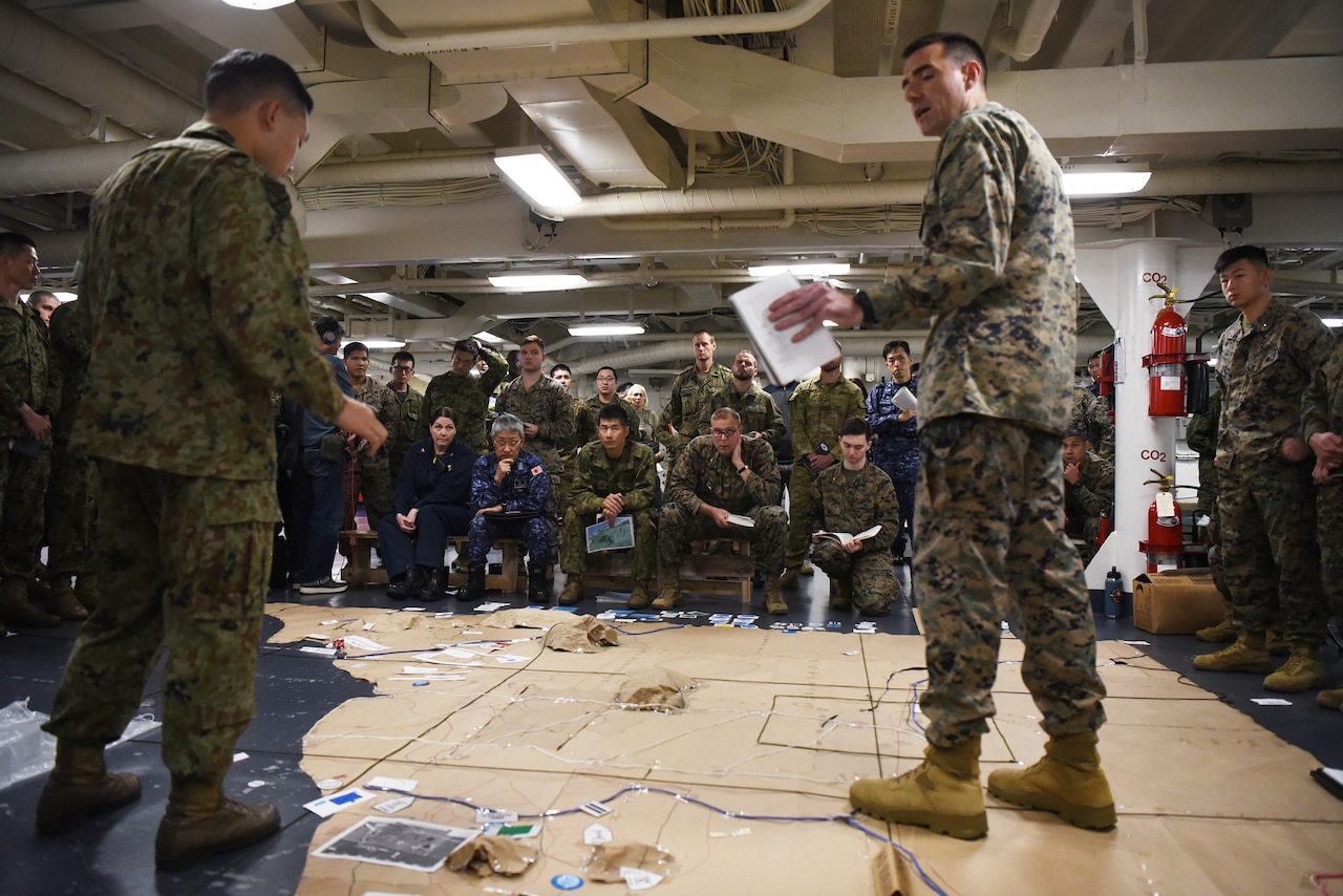A large group of service members sit inside and look at a big map.