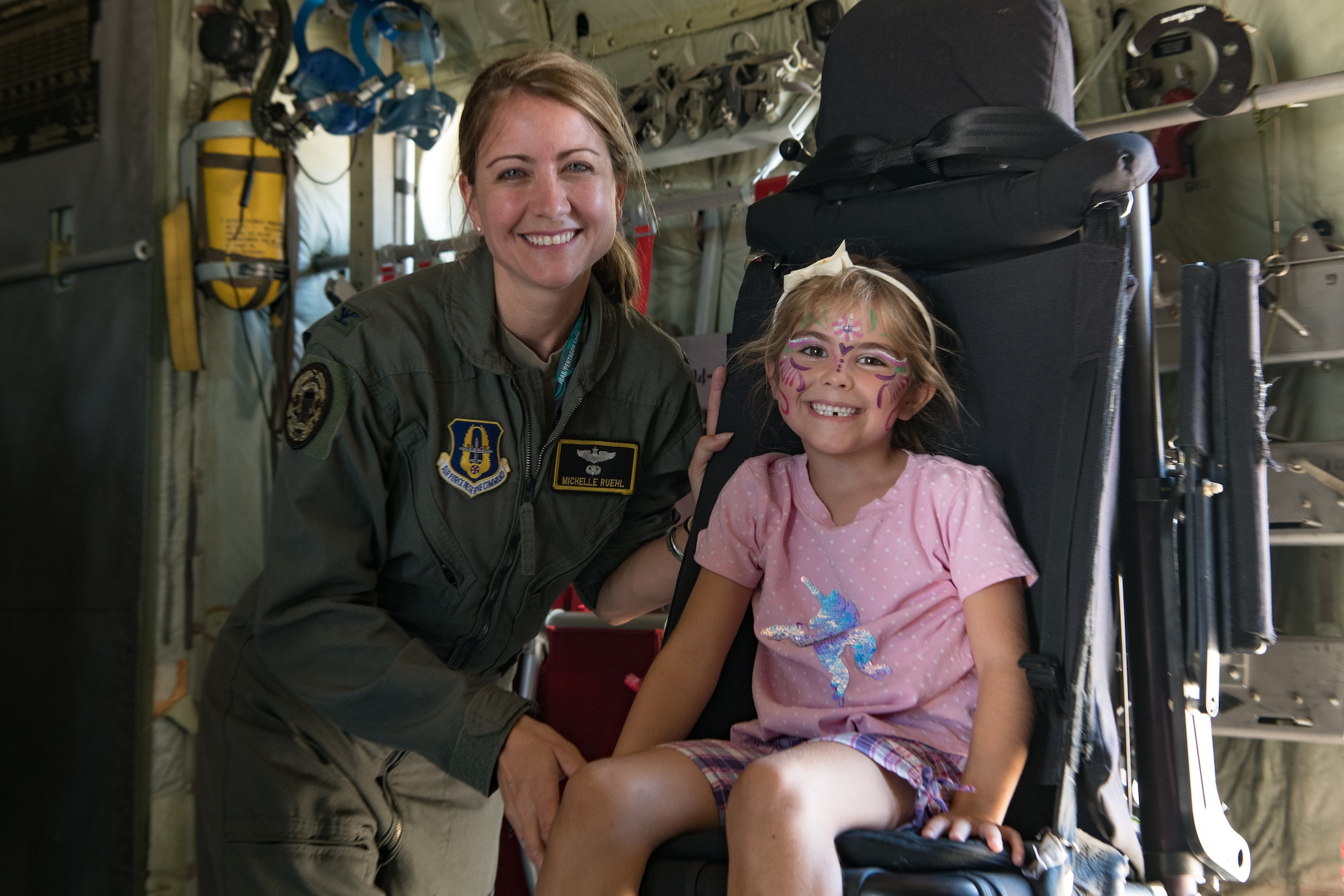 A woman in a flight suit kneels next to a child sitting in a black seat with paint on her face in the cargo bay of a C-130.