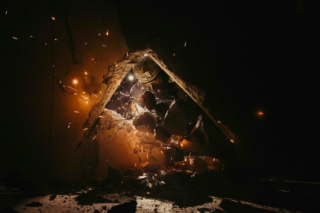 Sparks fly as a Marine wearing protective gear cuts concretes in the dark illuminated by dark orange light.