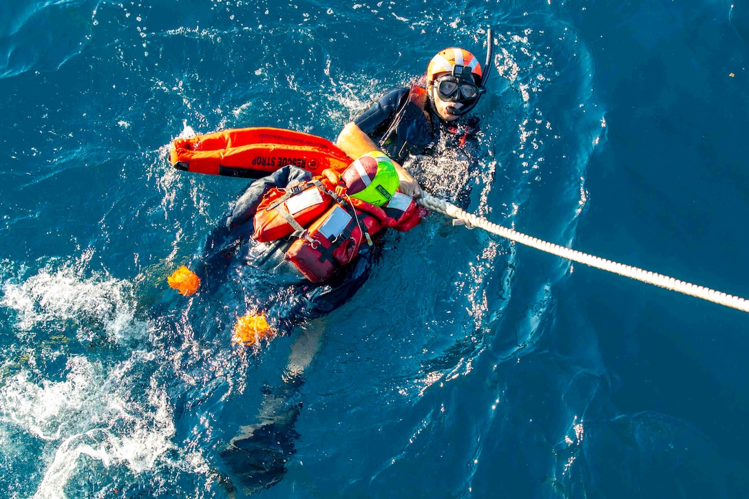 A sailor wearing swimming gear while floating on their back holds a dummy in a life vest and a rope.