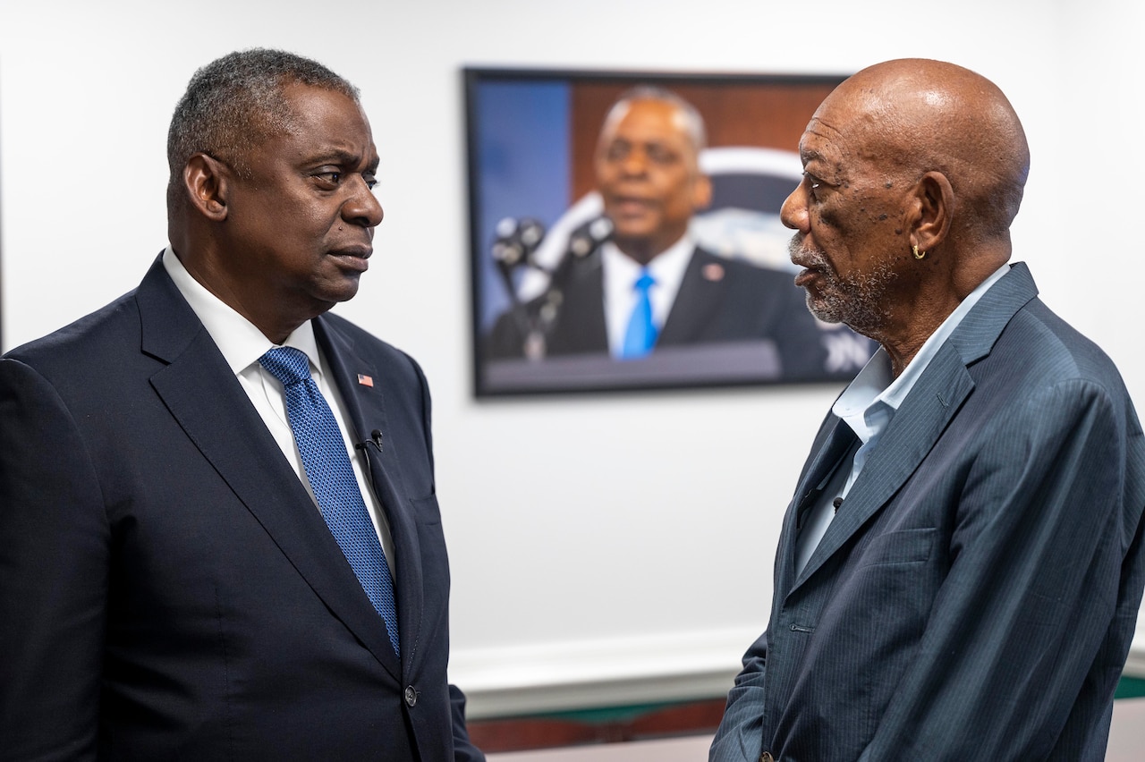Secretary of Defense Lloyd J. Austin III and actor Morgan Freeman talk in a hallway with Austin's photo in the background.