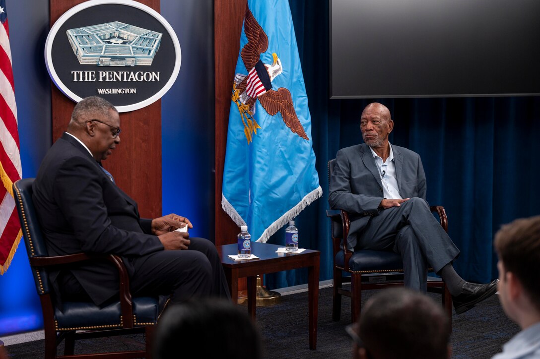 Secretary of Defense Lloyd J. Austin III and Morgan Freeman sit at a podium with the Pentagon logo and flags in the background.