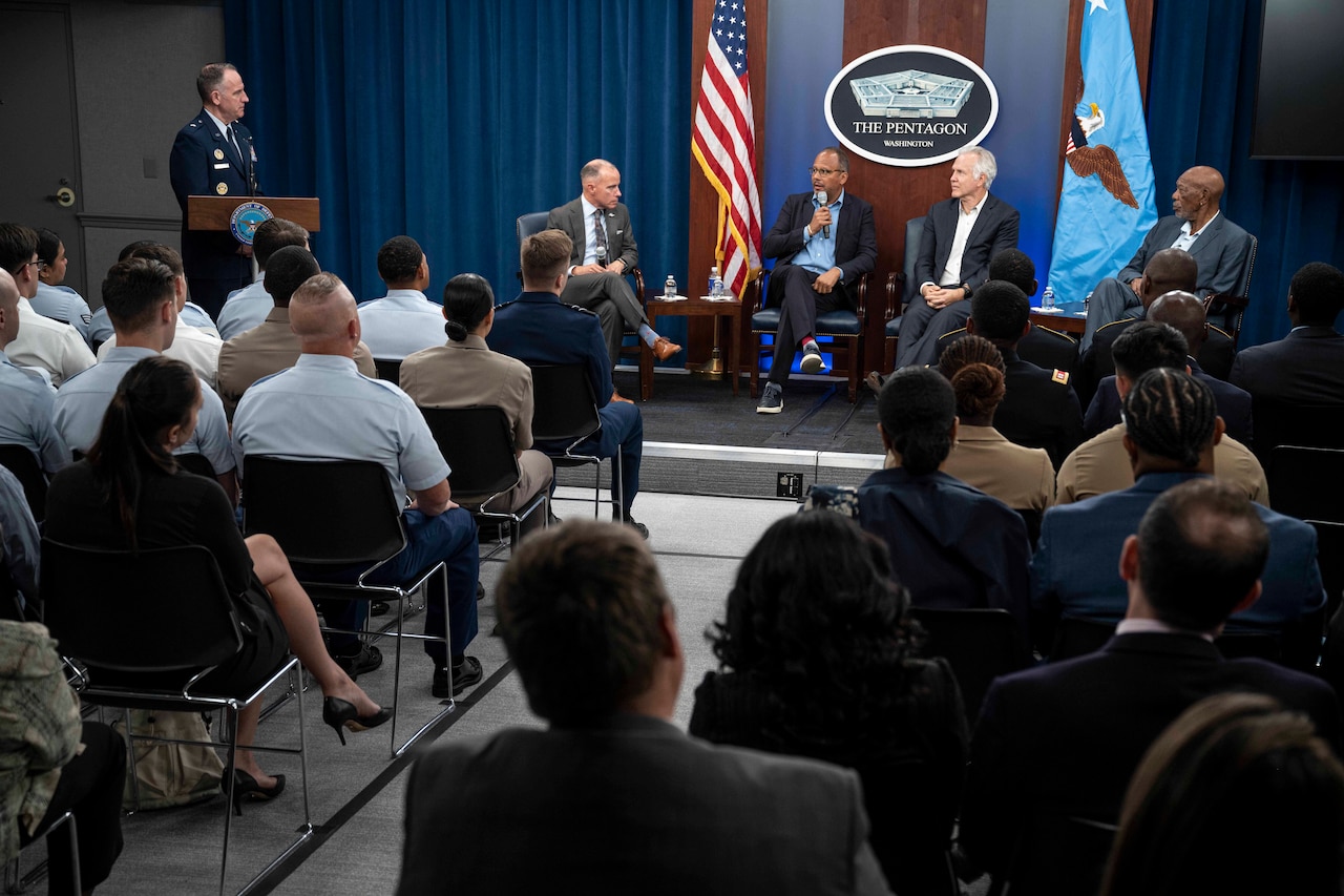 Air Force Brig. Gen. Pat Ryder stands at a lectern while Morgan Freeman and three others sit in discussion with an audience in the foreground.