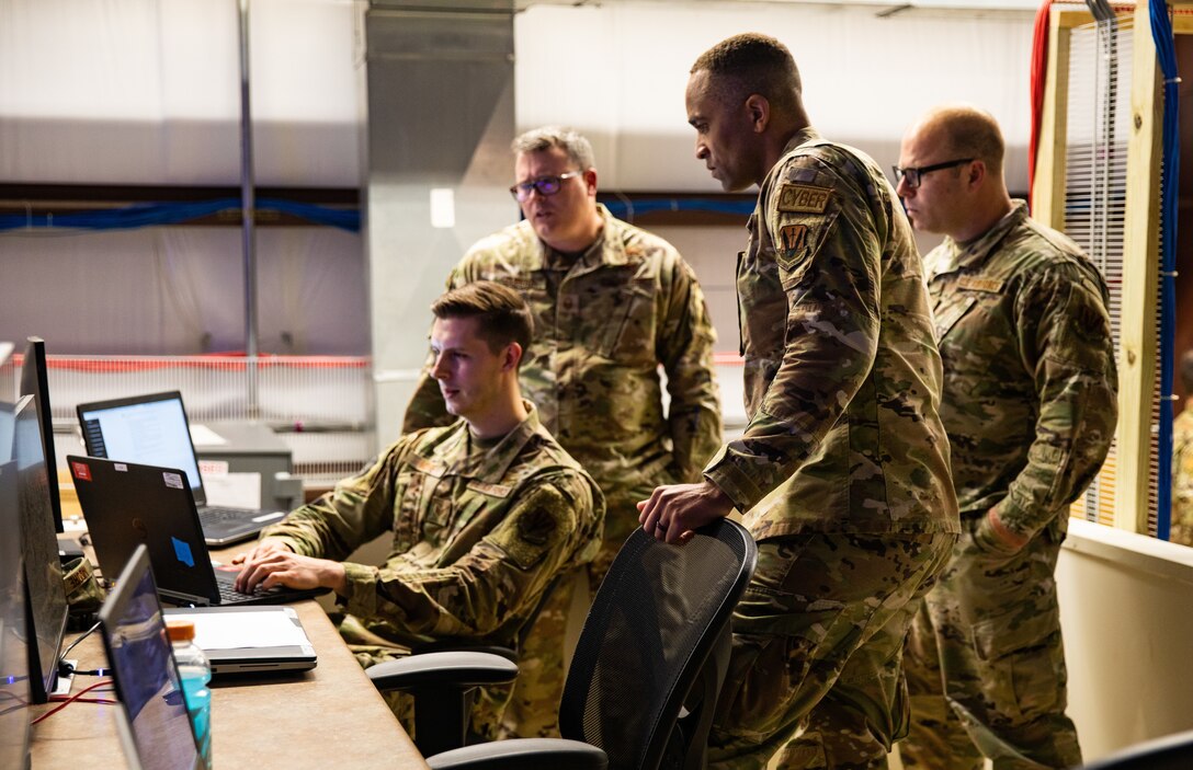 Service members in uniform huddle around a computer workstation.