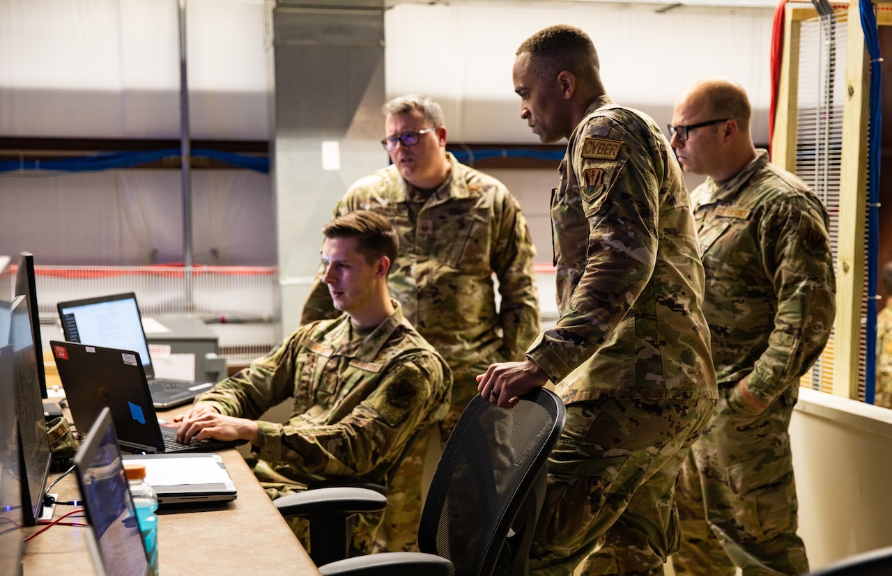 Service members in uniform huddle around a computer workstation.