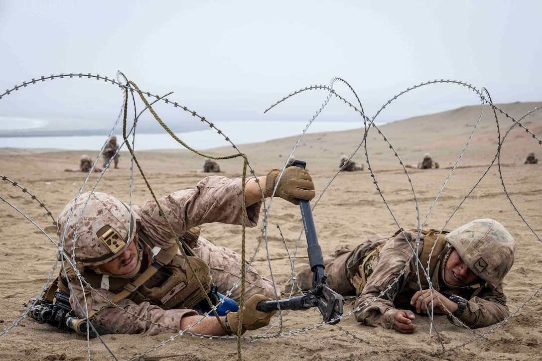 A Marine lying on the ground uses a bolt cutter to cut barbed wire as a service member lies beside.