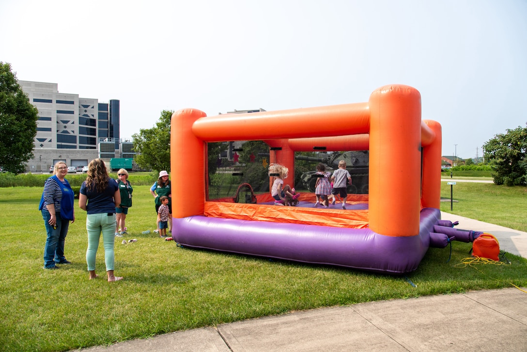 A large square bouncy house with purple bottom and orange top sits in a grassy area. many children bounce inside. Adults stand around watching from the outside.